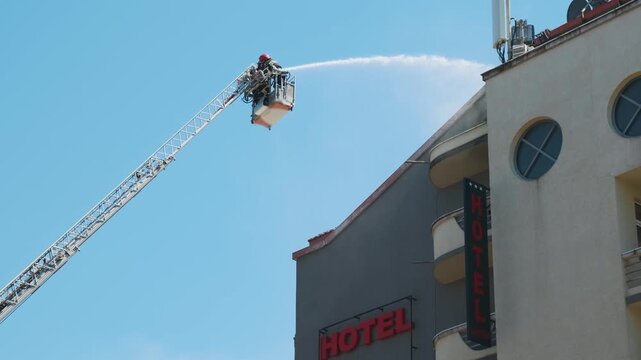 Firefighters in elevated platform extinguish burning house using extended ladder equipped with water hose to spray water onto the building roof with smoke. Fireman extinguishing fire in the city
