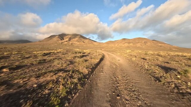 Flying low over dirt road and desert with mountains in background. FPV drone point of view, cinematic birds eye view with blue sky with white clouds.