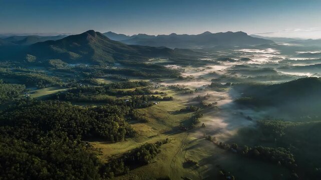 Aerial view of rolling green fields and misty valleys with a distant mountain range at dawn; warm sunrise light paints the landscape as fog drifts across the plains