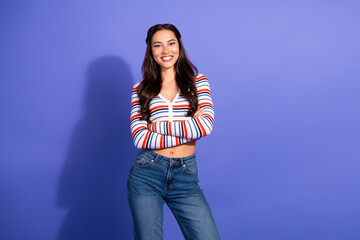 Confident young woman in striped top and jeans posing against purple backdrop smiling