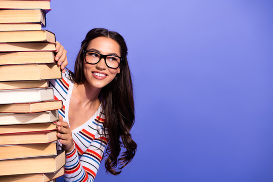 Young brunette woman with glasses holding a tall stack of books smiling against a purple background for education fashion lifestyle advertising and college campus stock photo