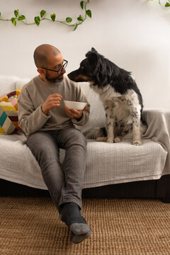 Adult man sitting on couch holding food plate while dog looks at him at home