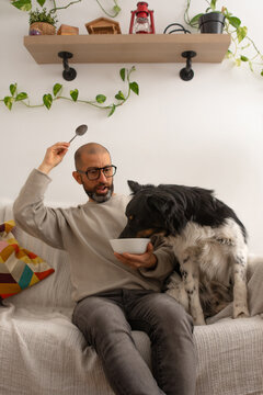 Adult man sitting on couch surprised as dog eats from his plate at home