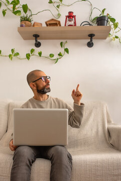 Adult man sitting on couch with laptop pointing to side at home vertical