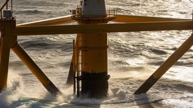 Crashing wave striking yellow column diagonal braces ladder at open sea sending spray by wind