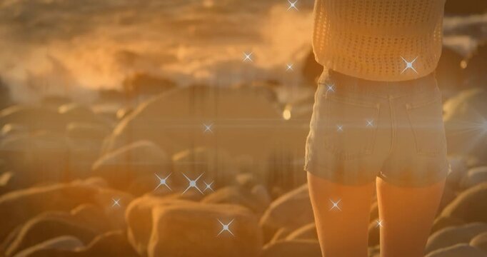 Animation of shining star icons over rear view of caucasian woman on the rocks near the sea