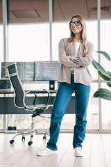 Confident female programmer standing in a modern office workspace with crossed arms in front of...