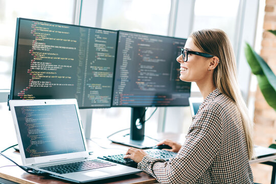 Female software developer coding at a modern multi-monitor workspace, smiling while working on laptop and desktop screens in a bright office environment