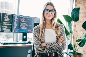 Naklejka premium Smiling female software developer in modern office with dual monitors coding at desk, confident young programmer wearing glasses and casual shirt in a tech startup workspace