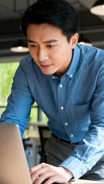 Vertical video: Entering office, male in blue shirt leaning at laptop, typing, checking charts