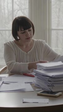 Vertical video: Seeing papers woman in top stacking them, sorting at table by window with red pen