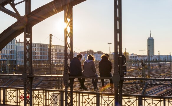 Three youths sitting on the balustrade of the Hackerbr&uuml;cke bridge over the railway tracks looking into the distance, back light, Munich, Upper Bavaria, Bavaria, Germany
