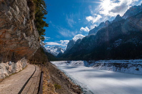 Hiking trail along frozen lake with rock overhang and alpine mountains at Gosausee Austria