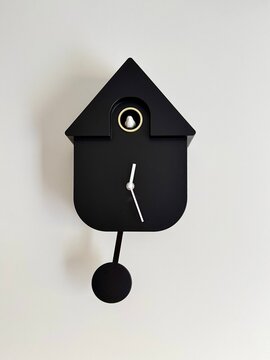 Black cuckoo clock with a minimalist design against a white wall. Features a triangular top resembling a house roof, a circular opening for the cuckoo, and simple white clock hands. 