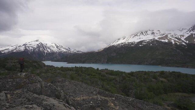 Hiker with a backpack resisting intense wind while overlooking a glacial lake and snowy mountains in nature