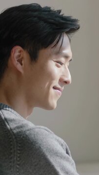 Vertical video: Smiling Asian man responding to soft light by pale wall in grey sweater, copy space