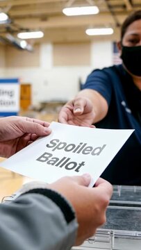 Handing Spoiled Ballot sheet toward clear ballot box in gymnasium, masked worker in navy polo