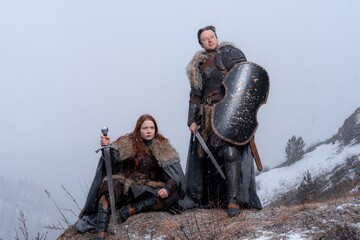 Naklejka premium A fierce warrior woman and a rugged man stand ready with swords and shields against a snowy mountain backdrop.
