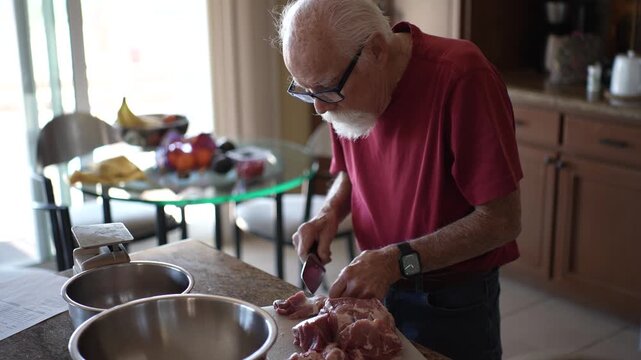Senior man slices meat in his home kitchen, working with a knife to prepare his meal at dinner time.