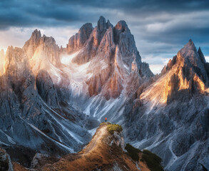 Fototapeta premium Girl on the mountain peak and high rocks at colorful sunset in autumn. Tre Cime, Dolomites, Italy. Colorful landscape with woman on trail, cliffs, grass, cloudy sky in fall. Hiking. Aerial view
