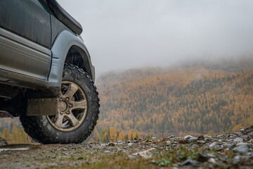 Close up of off-road 4x4 vehicle wheel on a wet gravel road against misty autumn forest. Expedition, adventure travel and overlanding concept in rainy weather. © ANDREY