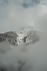 Obraz premium Dense white fog drifts through a rugged alpine valley, partially veiling the massive, snow-covered mountain faces under a heavy, overcast sky.