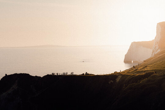 View of Durdle Door's chalk cliffs meet the tranquil sea under a hazy sky, where tiny figures stand silhouetted against the horizon, Wareham, Dorset, United Kingdom.