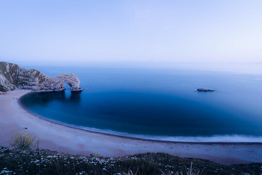View of Durdle Door's majestic limestone arch embracing the tranquil, deep-blue sea under the soft, ethereal glow of twilight, Wareham, dorset, United Kingdom.