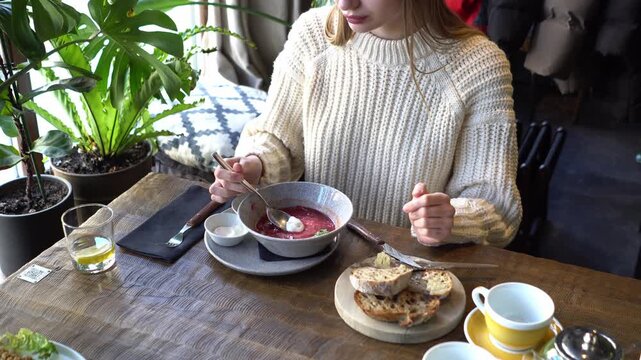 young beautiful woman eats borscht with sour cream and bread at cafe, girl adds sour cream to Ukrainian dish and stirs it in restaurant