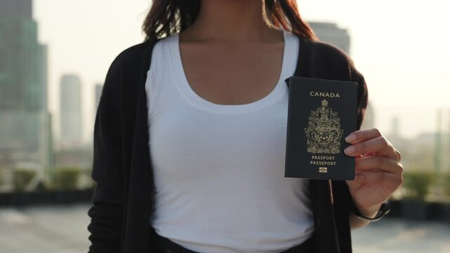 Crop view of woman hand holds passport of Canada against skyscrapers silhouettes in city. Female shows proudness of Canadian citizenship with identification document outdoors