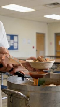 Vertical video: Service start, server in apron ladling stew into bowl and stirring pot at cafeteria