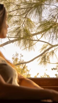 Woman sitting wooden seat, warm sunlight through pines causing leaning back, relaxing for wellness
