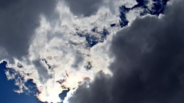Dramatic sky with dark clouds swirling over bright blue. Sunlight breaks through, illuminating fluffy white edges. Moody atmosphere perfect for cinematic projects