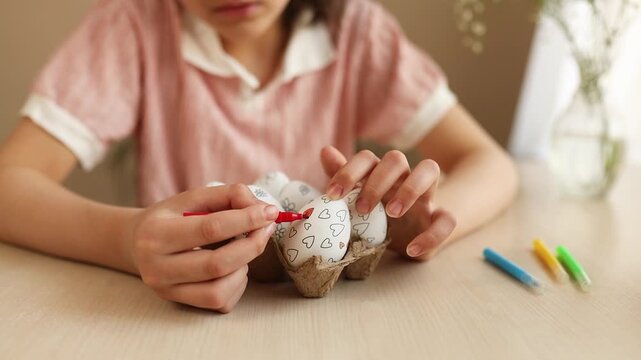Close-up of girl's hands decorating Easter eggs with red marker. White eggs with hand-drawn hearts and flowers placed in egg carton on table. Creative Easter preparation, DIY holiday decoration and sp