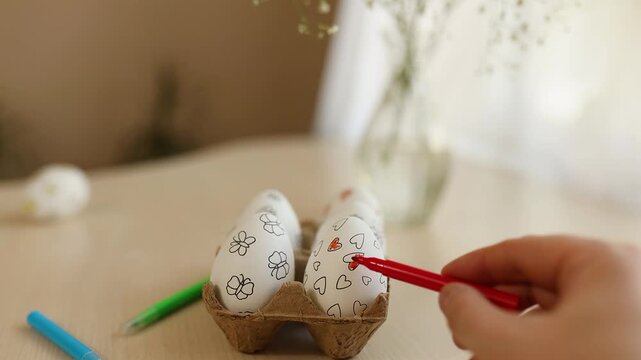 Close-up woman's hand decorating Easter eggs with red marker. White eggs with hand-drawn hearts and flowers placed in an egg carton on table. Creative Easter preparation, DIY holiday decoration and sp