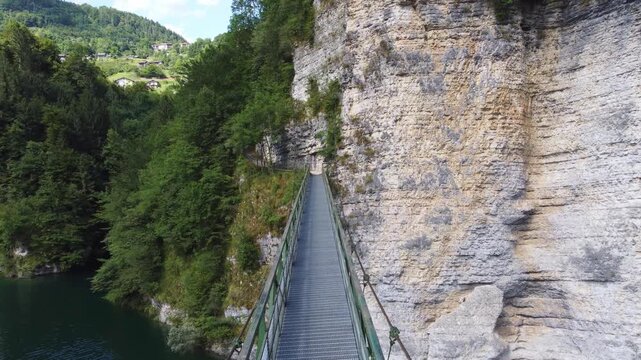 Aerial shot following a man walking across a suspension footbridge over Lake Senaiga, Italy. The camera moves towards a dramatic rocky cliff face and a path carved into the mountain.