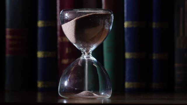 3 minute hourglass starting to flow, bookshelf background. Shallow depth of field, gold embossed letters on old books in low light, centrally framed with copy space on both sides
