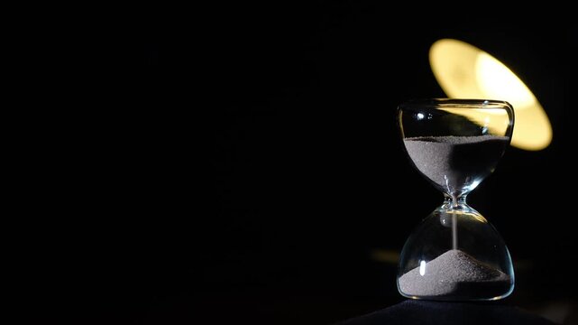 3 minute hourglass approaching end on black background. Soft yellow reading lamp bokeh behind upper glass, right aligned with extreme copy space, cinematic low light, 3 min sand timer
