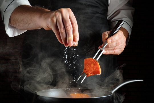 A chef is focused on cooking in a kitchen. He holds a fork with a piece of food while pouring salt from his other hand. Steam rises from the frying pan as he works