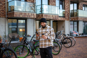 Obraz premium Young man commuting with e-bike, drinking coffee and checking phone in a modern city neighborhood