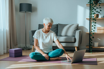 Fototapeta premium Middle aged woman sitting on yoga mat and using laptop during home workout session, representing online fitness learning