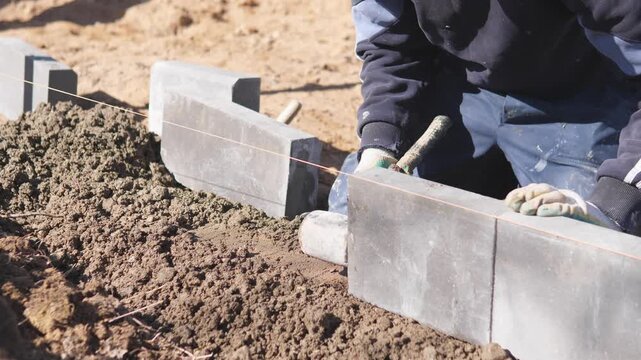 Construction Site Worker Laying Concrete Blocks Kneeling On Soil, Using String Line To Set Straight Row, Tapping Blocks Into Mortar With Mallet, Wearing Gloves And Work Jeans, Closeup Hands