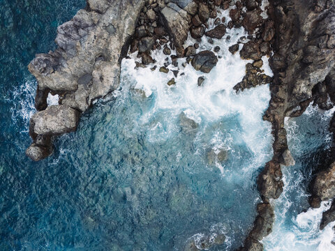 Aerial top-down view of turquoise ocean waves crashing against a rugged rocky shoreline. The dynamic movement of white sea foam contrasts with dark volcanic stones, creating a dramatic natural texture