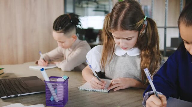 Students write in notebooks at a desk in a modern classroom during a study session in the afternoon with a laptop and stationery present