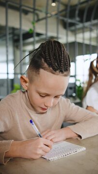 Boy writes in exercise book while seated at a table in a modern workspace during the daytime with another child nearby,vertical video