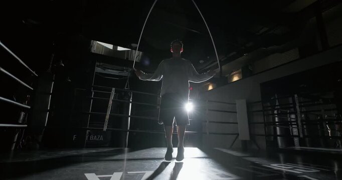 Man jumping rope during workout in a dark gym. Athlete training with skipping rope under dramatic lighting, silhouette view. Fitness and cardio exercise