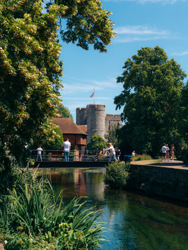 View of a serene river reflects the ancient stone castle under a bright sky, framed by lush green trees and vibrant foliage, Canterbury, England, United Kingdom.