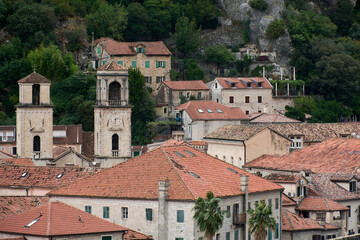 Fototapeta premium View of historic stone buildings and orange tile roofs in the old town of Kotor with a clock tower and church bell tower against a green hillside.