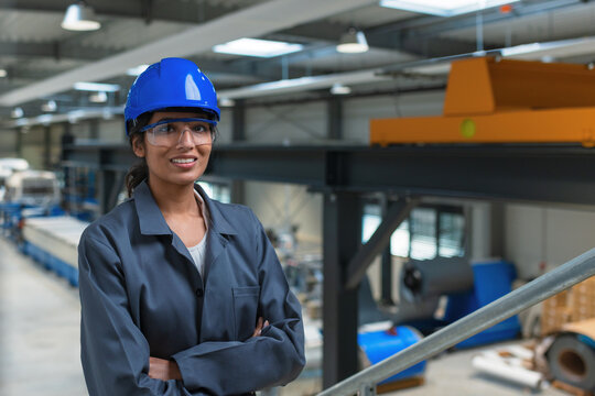 An Indian female engineer stands in a factory environment wearing safety gear and overseeing machinery operations on a production line.