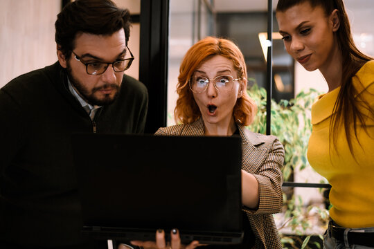Three colleagues gather around a laptop in a modern office, expressing surprise and excitement over a presentation or data on the screen during a midday work session